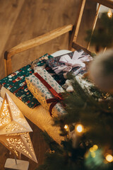 Stylish christmas gifts in festive wrapping paper with bows, vintage ornaments on chair on background of golden christmas star and tree lights. Happy holidays! Atmospheric christmas eve