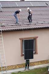Men technicians lifting up photovoltaic solar moduls on roof of house. Builders in helmets installing solar panel system outdoors. Concept of alternative and renewable energy.