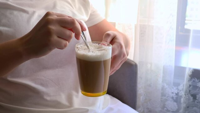 Woman hands mixing cappuccino coffee in a glass cup with a spoon in the morning close up