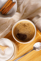 Cup of black coffe served on a white mug. There is a saucer with white sugar on a wooden tray, and everything lays on a white tablecloth. Upside view
