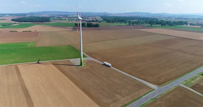 A white truck drives along the fields next to the wind turbine. The truck is driving down a steep road. A white truck delivers cargo along a beautiful road.