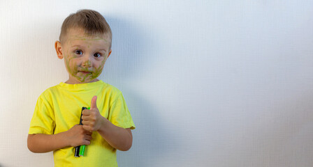 A 3-year-old boy painted his face with a marker. The boy stands against a white wall, holding markers in his hands, smiling and showing the class. Banner with space for text