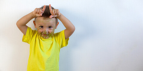 A 3-year-old boy painted his face with a marker. The boy stands at the white wall and makes horns....