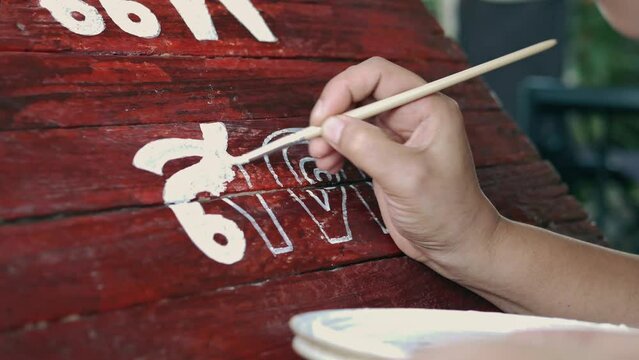 Close-up of Hands of Young woman painter creating art use a paintbrush to draw white lettering designs on a wooden coffee shop sign. outdoor activities, People doing activities.4k