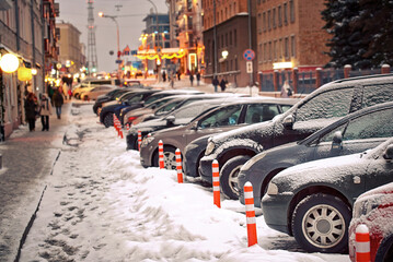 Cars parked close to flexible plastic warning post. Cars parked at parking lot along city road in winter season. Heavy snowfall or snowstorm in the city. Cars covered with snow - obrazy, fototapety, plakaty