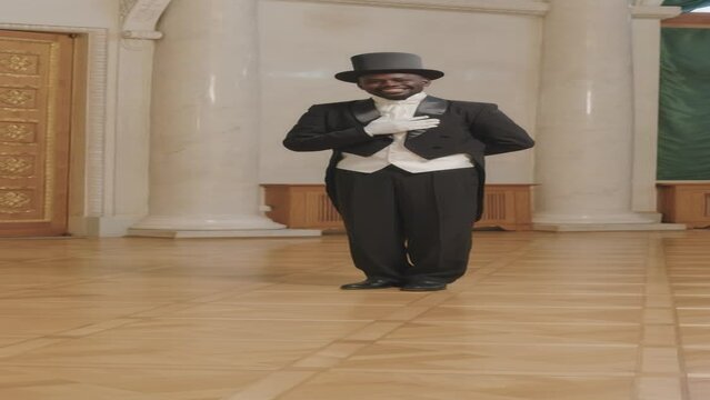 Vertical full length slowmo portrait of young Black gentleman wearing black tuxedo and cylinder hat bowing down with his hand on chest standing on parquet floor in elegant classic ballroom
