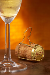 Champagne cork, Happy new year and 2024 text on golden cap. Glass in front, wooden table.
