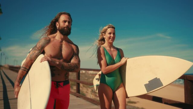 A couple of surfers on the beach. Caucasian woman in a swimsuit and a beefy bearded man with tattoos in swimming trunks holding surfboards walk along a wooden bridge to the sea.
