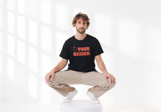 Mockup of man wearing customizable t-shirt in studio crouching on plinth