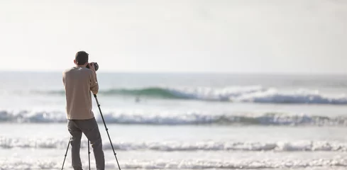 Crédence de cuisine en verre imprimé Coucher de soleil sur la plage Young handsome photographer with a tripod and a camera on the beach