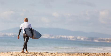 Fototapeta na wymiar Surfing the Waves: A Man Carrying a Surfboard on a Serene, Sunlit Sandy Beach