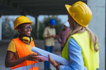 Two workers talking at a construction site, standing in a building in the framing stage. The focus is on the African American woman, wearing a yellow hard hat, holding a set of rolled-up plans