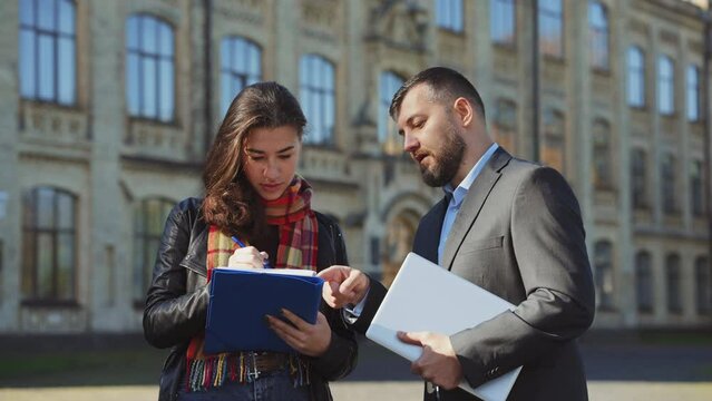 Female student taking notes in her project work, male mentor assisting her. Professor tutoring student with homework or thesis. Blurred campus background. Concept of education