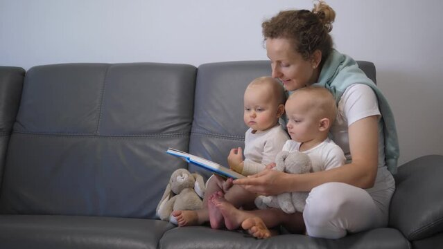 Mother and kids sit on sofa. Wide shot of mother reading book for kids, children touch picture with fingers. Concept of reading books for develop kids imagination and brings family closer together.