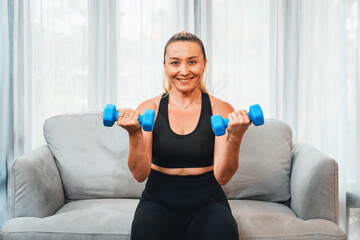 Fototapeta na wymiar Athletic and sporty senior woman engaging in body workout routine, sitting on sofa and lifting dumbbell at home as concept of healthy fit body with body weight lifestyle after retirement. Clout
