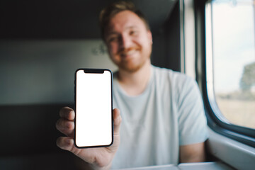 A man with a beard and mustache in a blue t-shirt is using a smartphone while traveling by Railway train, sitting in the train and looking out the window.