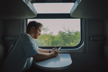 A man with a beard and mustache in a blue t-shirt is using a smartphone while traveling by Railway train, sitting in the train and looking out the window.