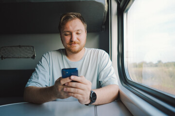 A man with a beard and mustache in a blue t-shirt is using a smartphone while traveling by Railway train, sitting in the train and looking out the window.