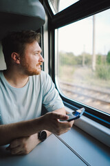 A man with a beard and mustache in a blue t-shirt is using a smartphone while traveling by Railway train, sitting in the train and looking out the window. - obrazy, fototapety, plakaty