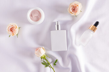 White matte bottles and a jar of cosmetic self-care cream on white satin with rosebuds. Top view....