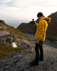 Man in yellow jacket filming video on a mirrorless camera during a hike in Lofoten, Norway.