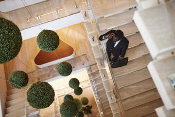 Top-down shot of busy African American businessman with leather bag wearing suit calling by phone standing on stairs in modern office building, copy space