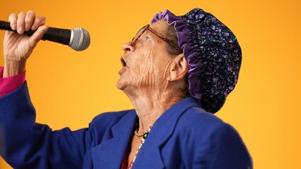 Closeup funny crazy toothless grandmother with a fashionable look with glasses, singing enthusiastically into a microphone and dancing isolated on solid yellow background