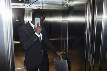 Medium long shot of Black man wearing business suit while talking on phone in office elevator and pressing lift button, copy space © Seventyfour