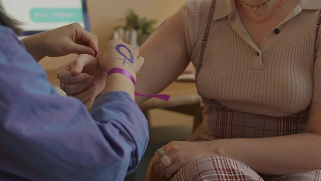 Cropped shot of two unrecognizable feminist activists tying up purple ribbon on wrists as symbol of protest and woman empowerment