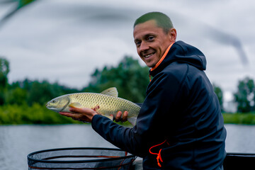 portrait of a professional fisherman holding a carp fish on the bank of a river fishing in...