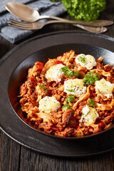 Lasagnette with beef ragu, mushrooms and ricotta cheese in black bowl on dark wooden table with fork and spoon at background, italian recipe, vertical view