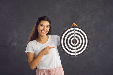 Studio portrait of young happy woman pointing finger at darts board she is holding in her hands....
