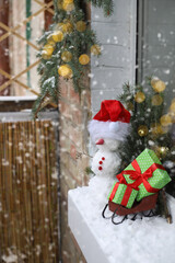 a snowman in a Santa Claus hat stands on the balcony. next to Santa Claus's sleigh with gifts in green wrapping paper with a red ribbon. spruce branches around
