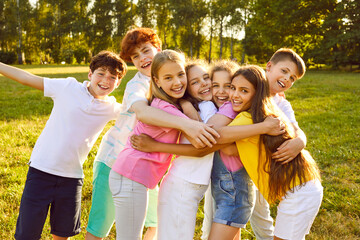 Children have fun and make friends at the summer camp. Group of kids spending time outdoors together. Bunch of happy girls and boys hugging each other in a green sunny park - obrazy, fototapety, plakaty