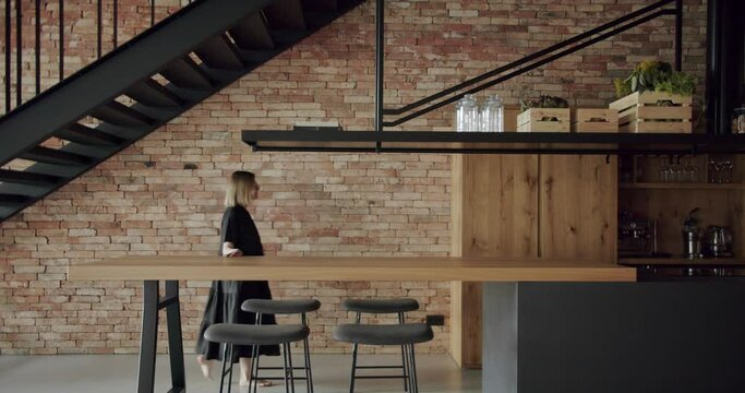 Woman walk near table with bar stools and touch wooden countertop at modern apartment. Cooking table and kitchen island at house with loft interior. Contemporary furniture at home with open plan