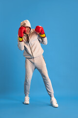 Young guy in grey sportswear standing in red boxing gloves, training against blue studio background. Fighter. Concept of human emotions, youth, sportive lifestyle, fashion, combat sport