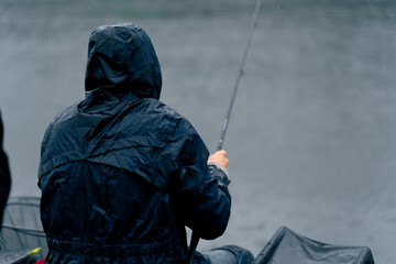 Fisherman in raincoat with fishing rod or spinning and professional tools sitting on river bank...