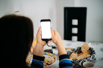 Woman from behind with dark hair looking at the phone screen in a family environment, with food in front of the table in the background.