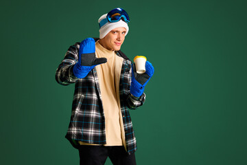 Hey. Young guy in warm jacket, gloves and hat standing with coffee cup and waving with hand against...