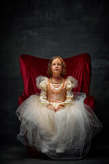Fototapeta premium Portrait of little girl, kid dressed like medieval person in old-fashioned dress sitting on throne against dark vintage background.