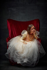 Portrait of boring girl, upset kid dressed like medieval person in old-fashioned dress sitting on throne against dark vintage background.