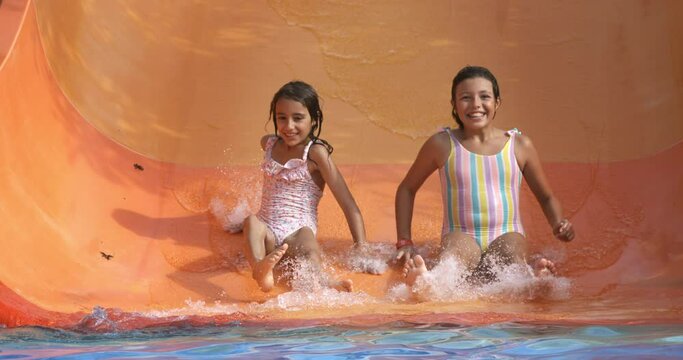 Super slow motion of two little girls friends or sisters in colorful swimsuits are having to slide down together at water slide in swimming pool at aqua park during summer holidays travel vacation.