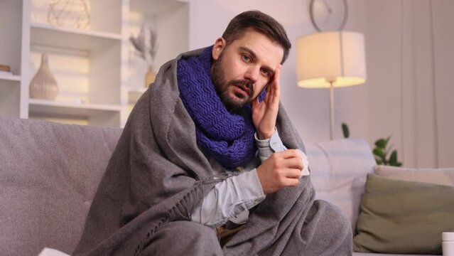 Sick unhealthy mature man on couch with warm scarf coughing sneezing and having severe headache temperature indoors Sad tired guy getting flu virus symptom at cold season and looking at camera at home