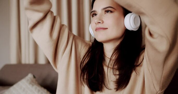 Mindful young happy woman wearing wireless headphones, listening relaxing music, resting on comfortable sofa, enjoying interesting audio book or affirmations, spending leisure indoors