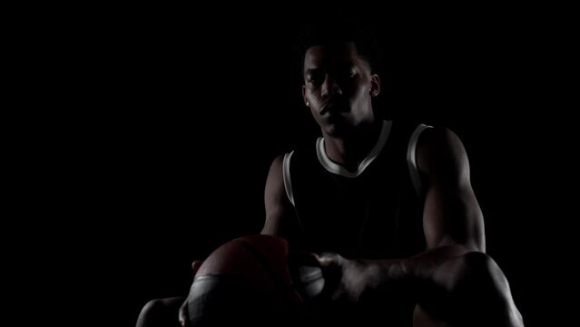 Basketball player sitting on a bench. Looking at camera, Concentrated african american sports man against black background.