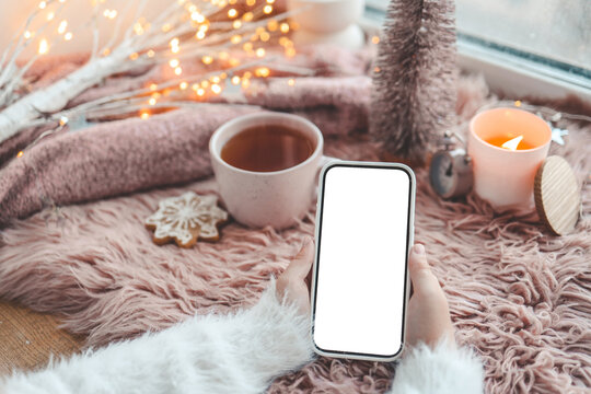 Phone in hands with an isolated screen against the background of a New Year's interior and a cup of tea