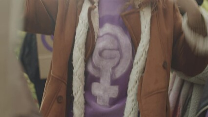 Tilting up shot of young Caucasian woman wearing purple t-shirt with feminist symbol raising hands and shouting while fighting for womens rights at outdoor protest with other female activists
