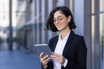 A smiling young businesswoman is standing near an office building and using a tablet. Talks on a video call, texts, close-up photos