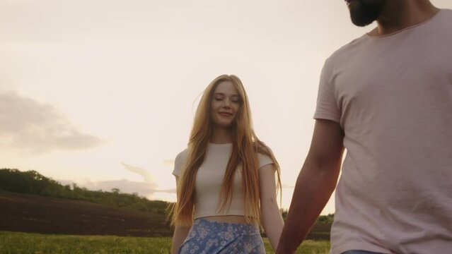 Handsome guy and red-haired woman walking on the field on a beautiful warm sunset, boy leading the girl, holding the hand. Concept about passion and love