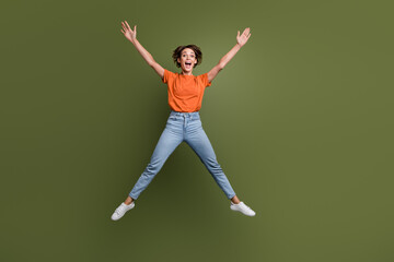 Fototapeta na wymiar Full size photo of funky optimistic woman dressed orange shirt jeans jumping hold raising arms up isolated on khaki color background